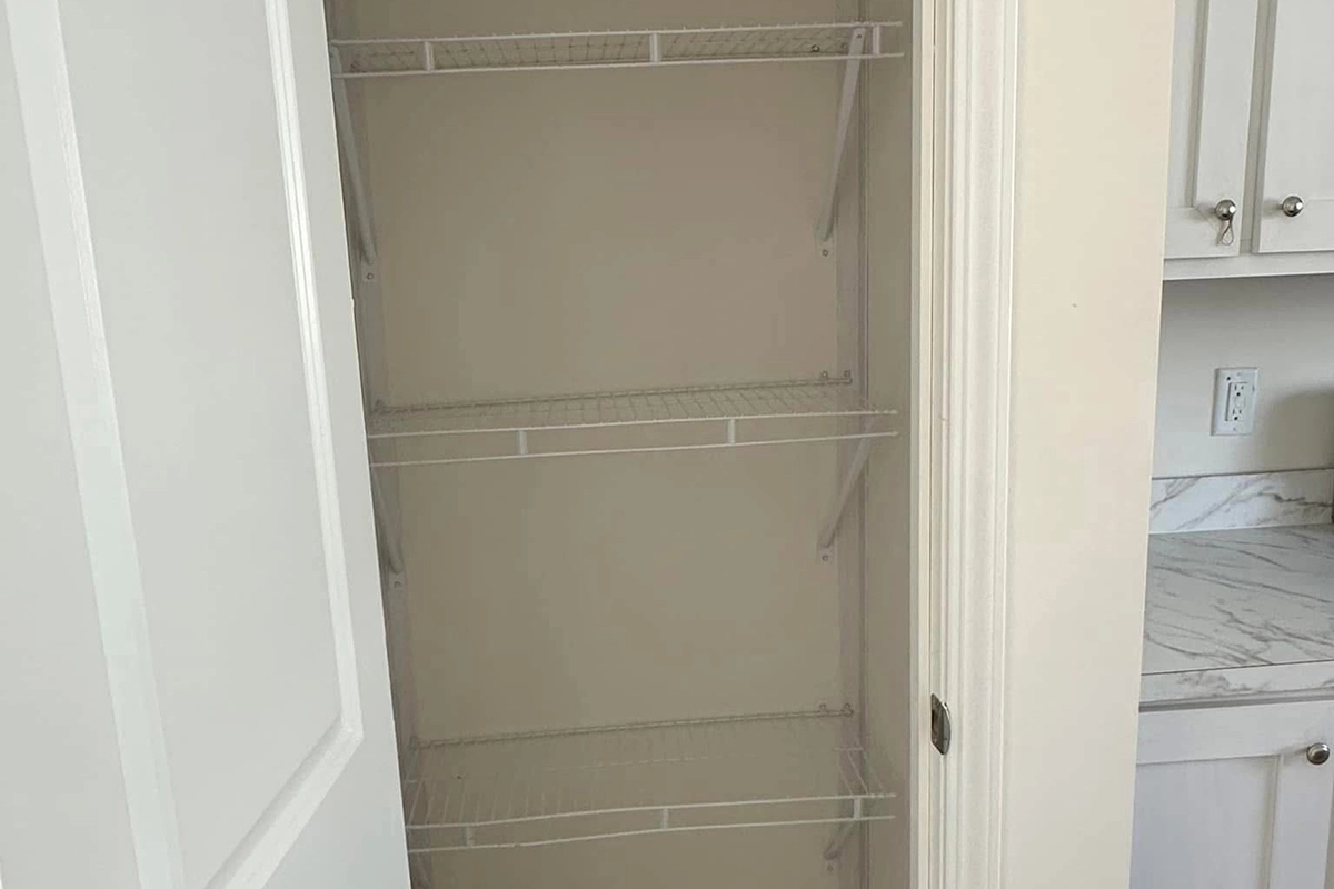 An empty closet with four white wire shelves, seen with the door open. The closet is next to a kitchen counter and white cabinets.