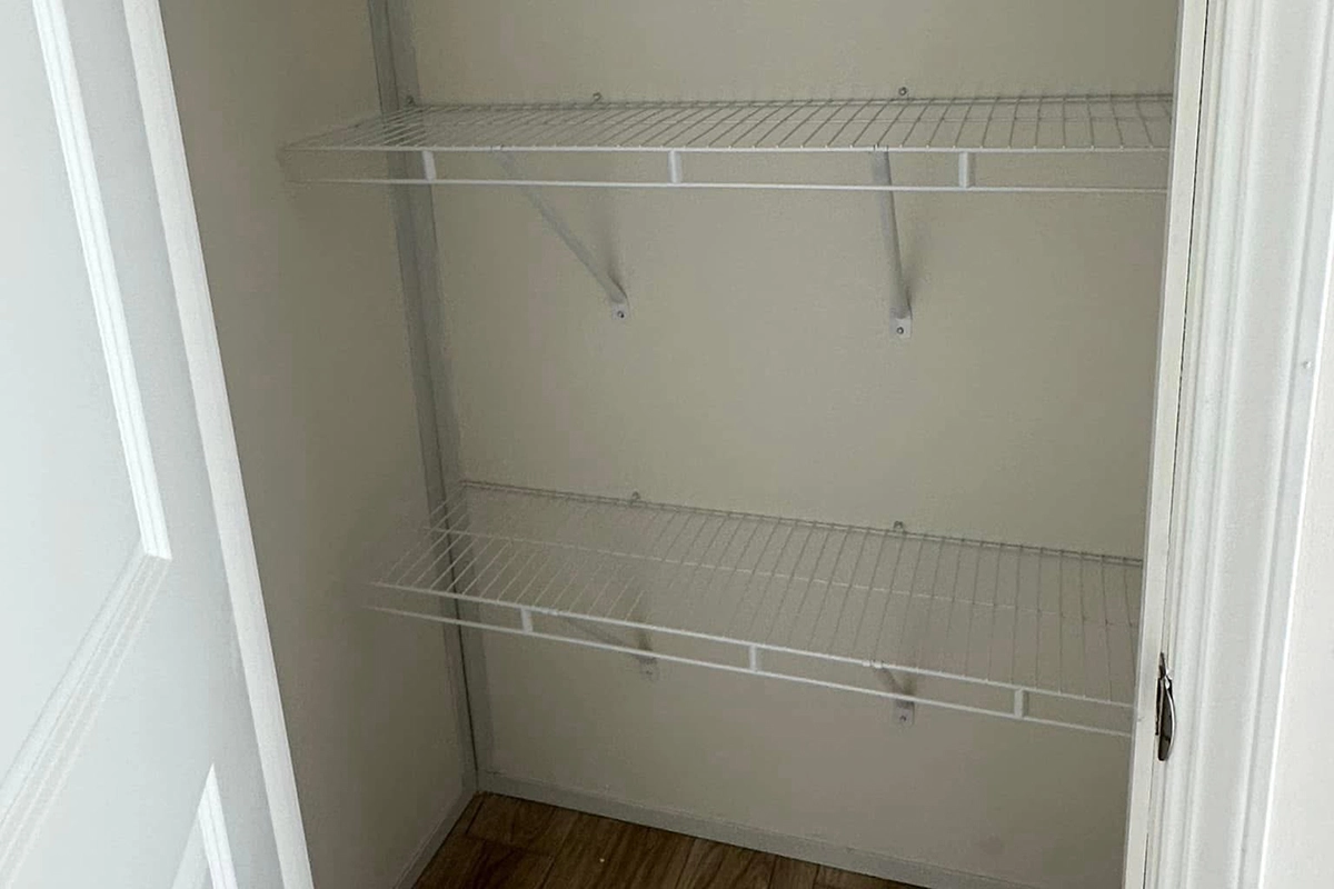 Empty closet with two white wire shelves mounted on the back wall, beige walls, and a wooden floor.