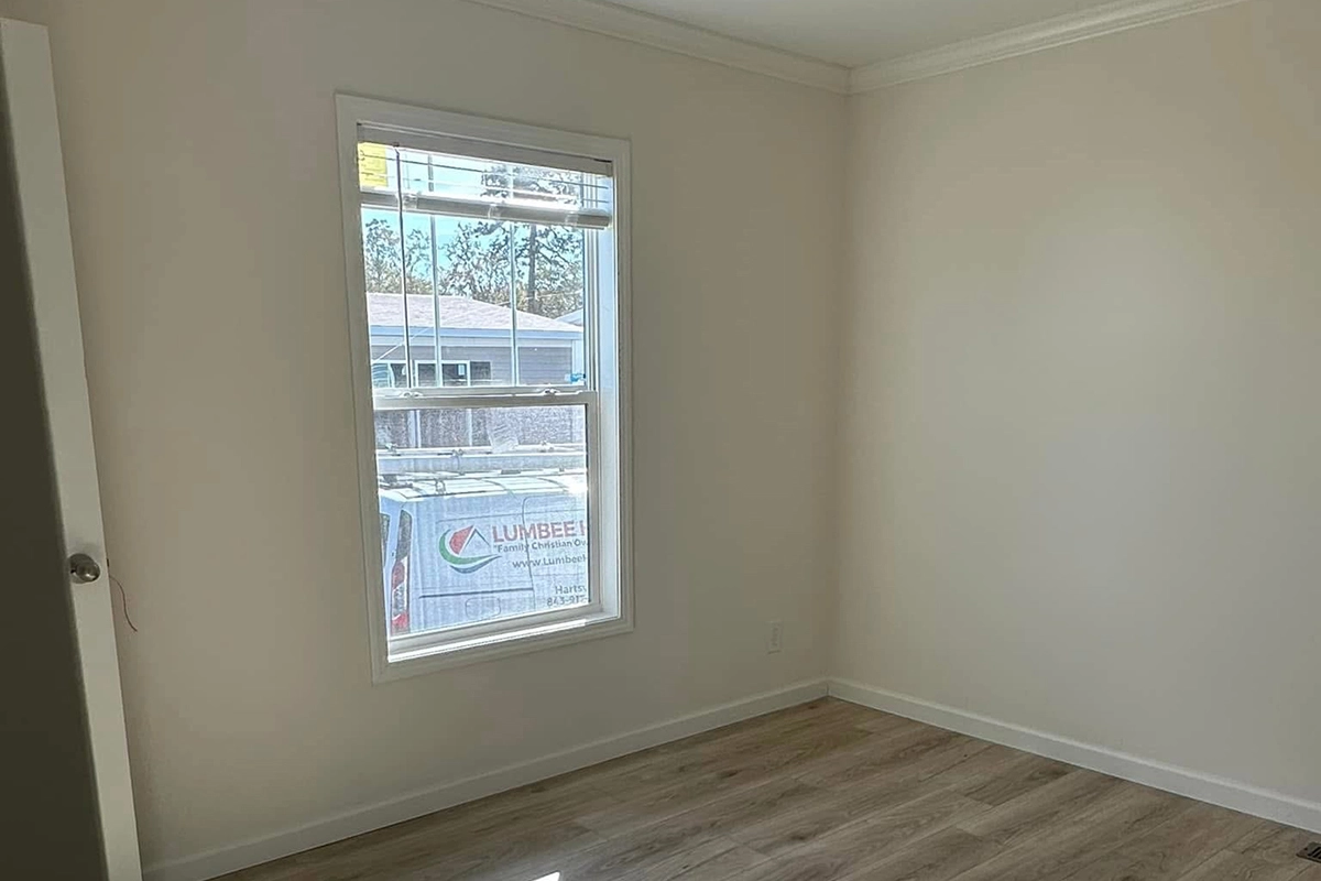 An empty room with light-colored walls, a window letting in daylight, and wood-patterned flooring. A building and a van are visible outside the window.
