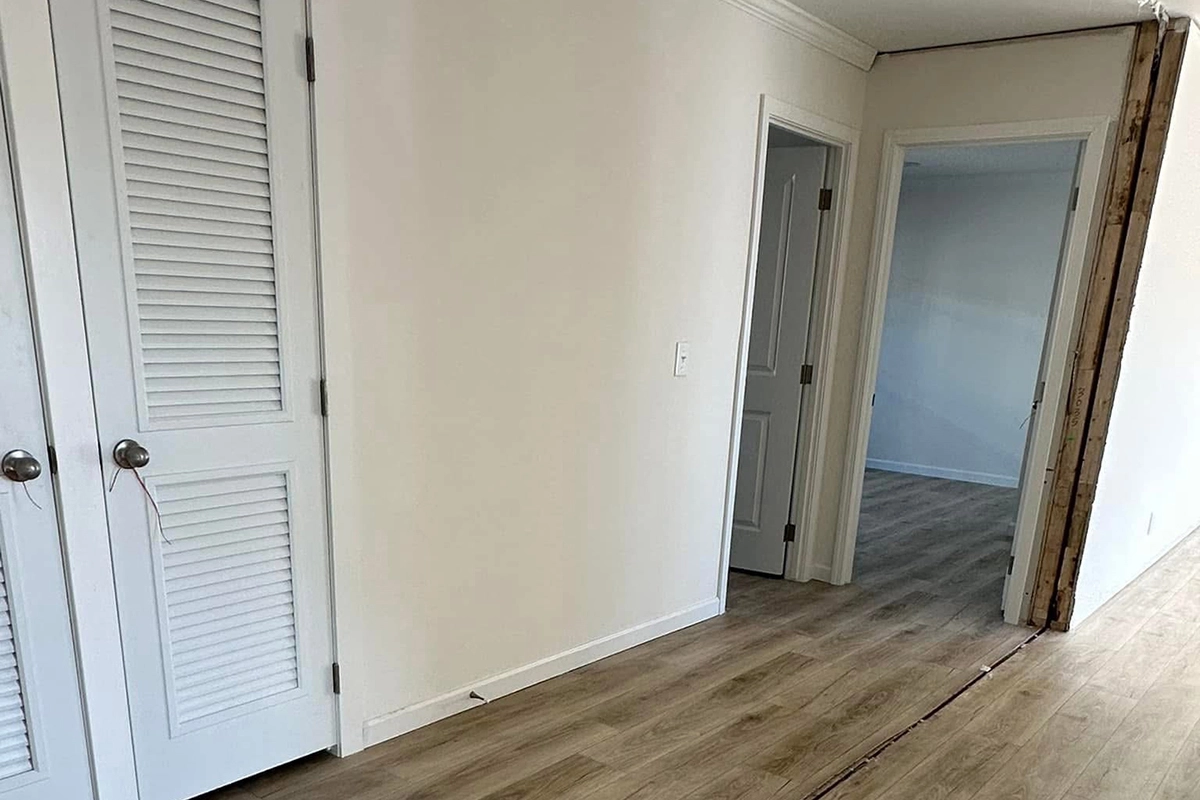 A hallway with light wood flooring, white doors on the left, and an open doorway leading to another room. Part of the wall on the right is unfinished.