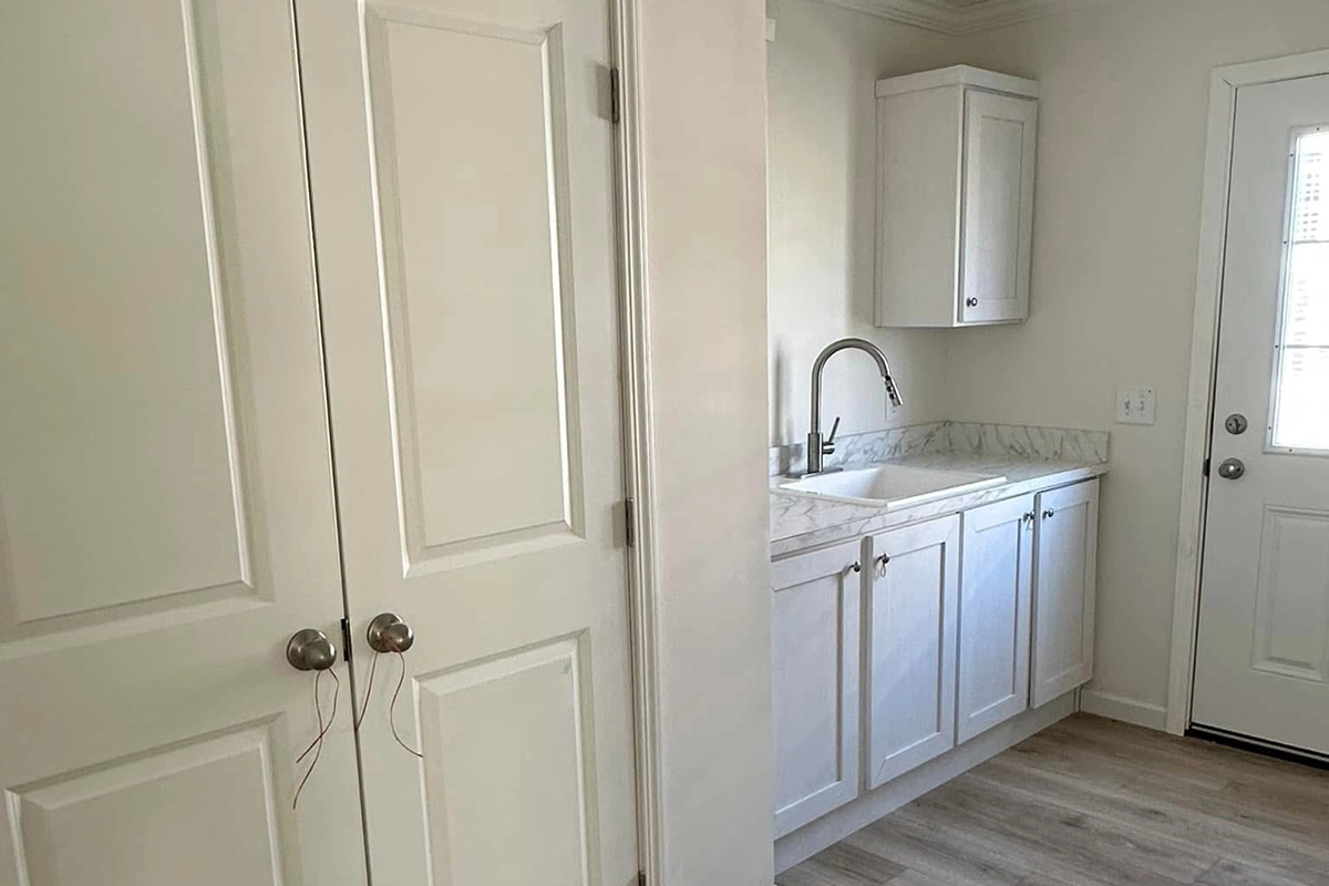 A small laundry room with white cabinets, a sink, marble countertop, double closet doors, and a door leading outside. Light wood flooring throughout.