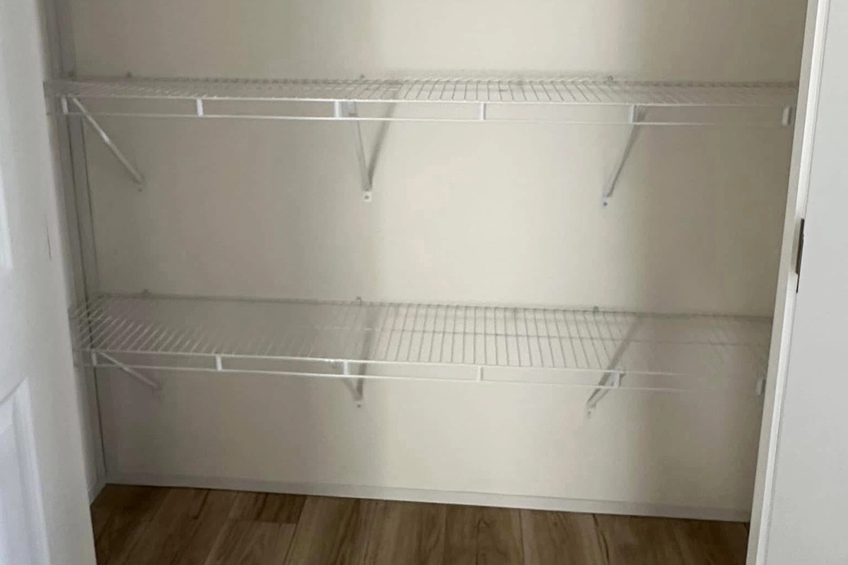 Empty closet with two white wire shelves mounted on a white wall, above a wooden floor.