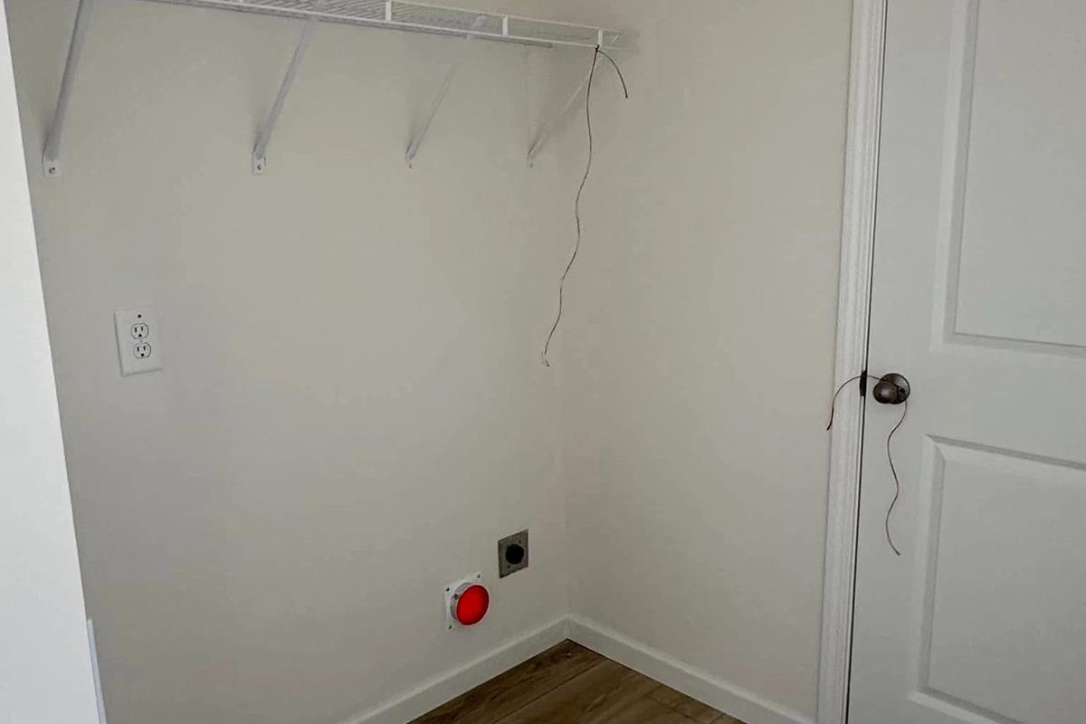 Empty laundry room corner with a white shelf, electrical outlet, wall vent, red circular fixture, and exposed wires near a closed door.