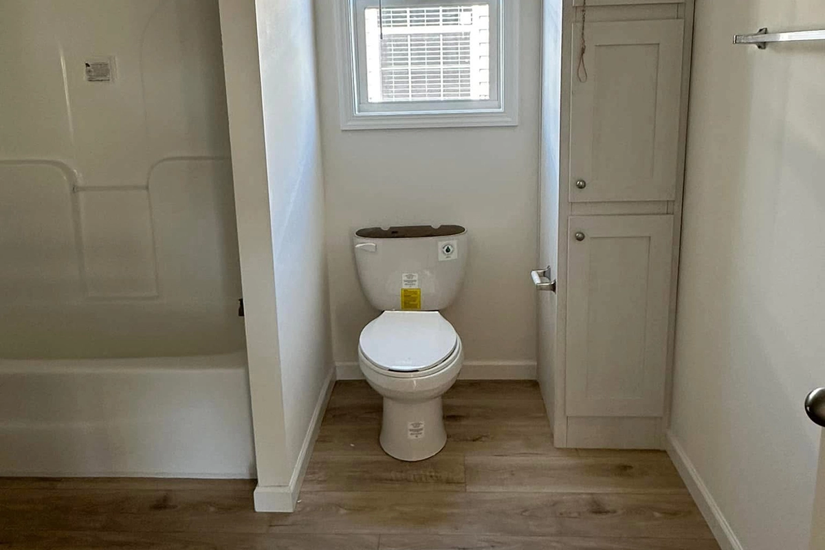 A small bathroom with a toilet under a window, a white cabinet to the right, a bathtub to the left, and wood-style flooring.