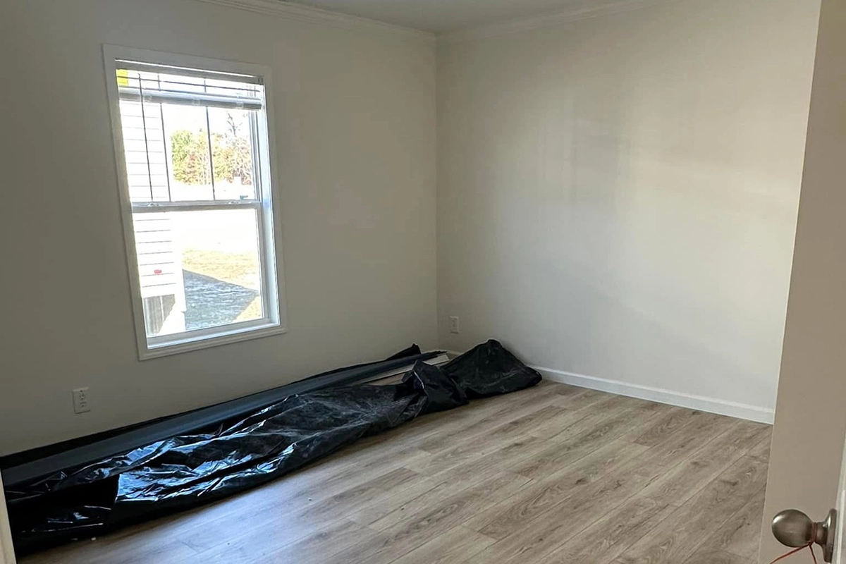Empty room with light-colored wood flooring, white walls, a window on the left, and a black plastic sheet lying on the floor along the wall.