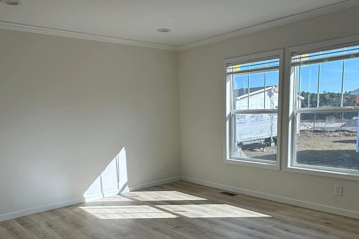 Empty, unfurnished room with light-colored walls and hardwood floor, sunlight streaming in through two large windows, construction visible outside.
