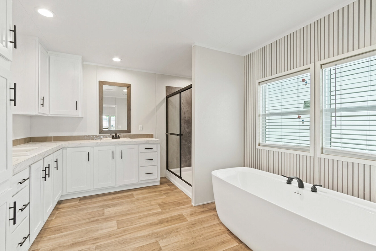 Modern bathroom with white cabinets, a large mirror above a sink, a freestanding bathtub, a glass-enclosed shower, and wood-look flooring. Two windows are on the right wall.