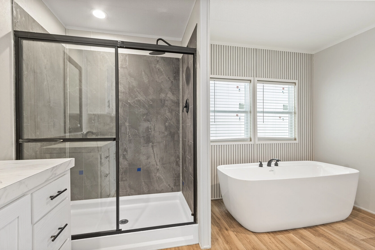 Modern bathroom with a glass-enclosed shower on the left and a freestanding white bathtub beneath two windows on the right, featuring wood-look flooring.