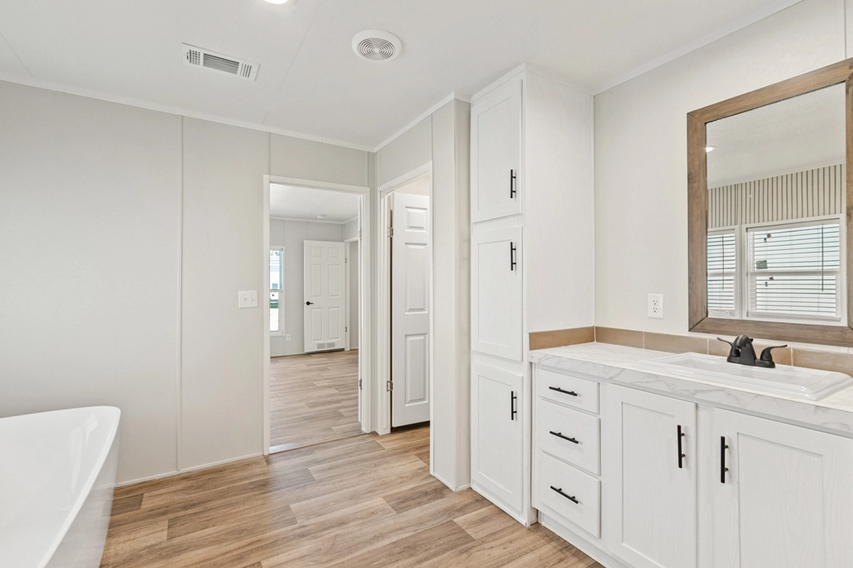 Modern bathroom with white cabinets, a single sink, a large mirror, and wood-style flooring. Doorways lead to other rooms with similar flooring and neutral walls.