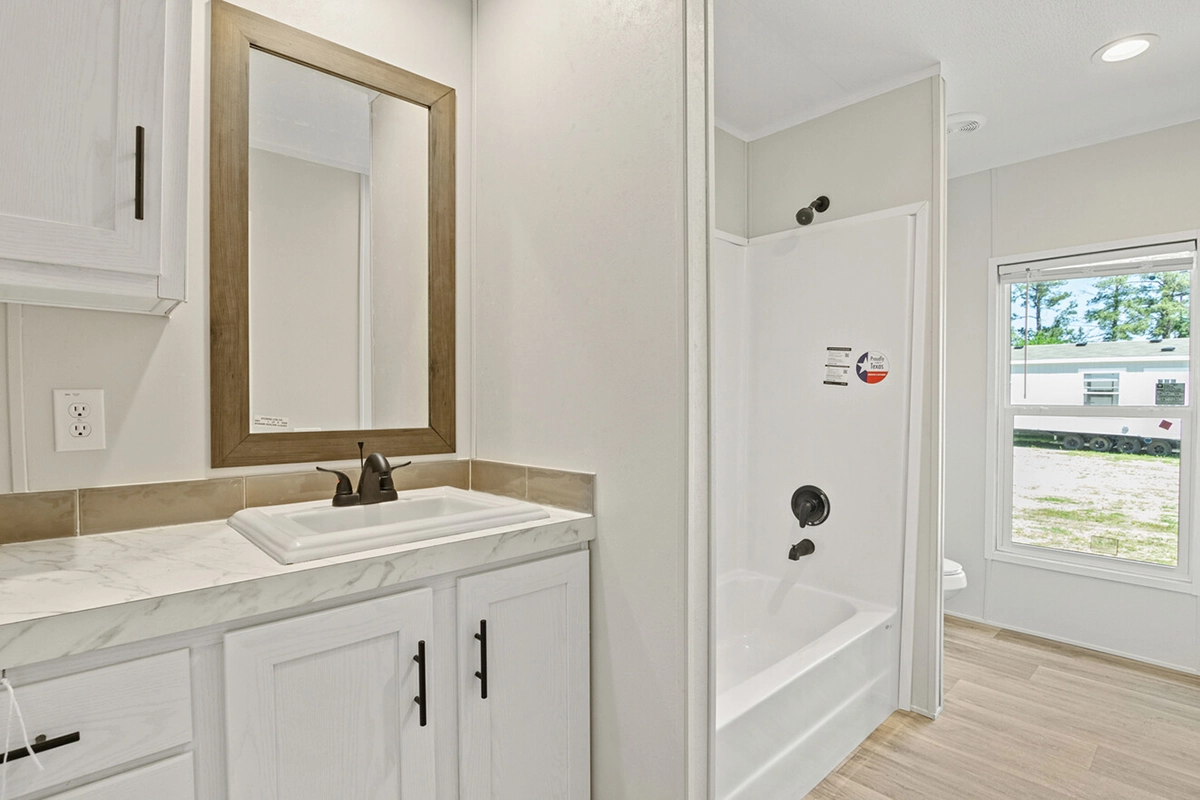 Modern bathroom with a white vanity and sink, brown framed mirror, bathtub with shower, and large window letting in natural light.