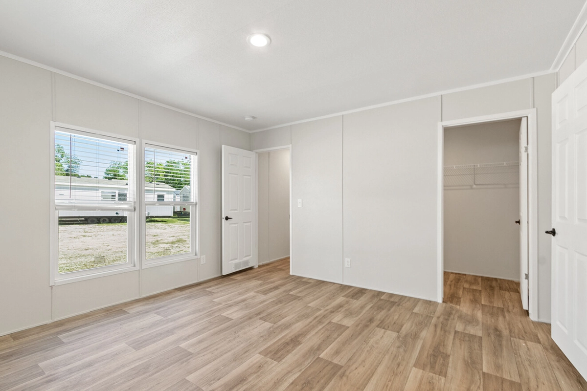 Empty room with light wood flooring, beige walls, two windows, two white doors, and a small walk-in closet with shelving.