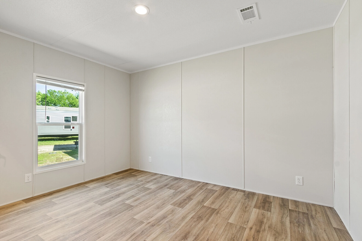 Empty room with light-colored walls, wood-style flooring, a single window letting in daylight, and a ceiling light fixture. Outlets and a vent are visible on the walls and ceiling.