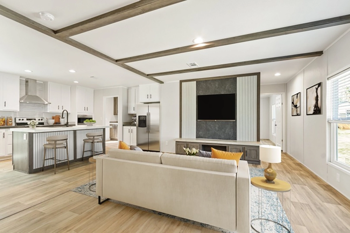 Modern open-plan living room and kitchen with neutral tones, a beige sofa, wall-mounted TV, bar stools at a kitchen island, and large windows letting in natural light.