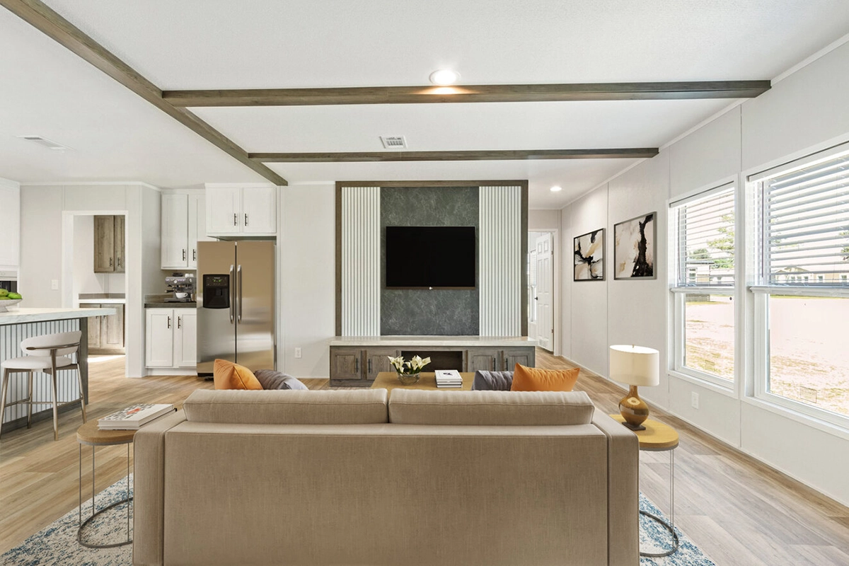 A modern living room with a beige sofa, wall-mounted TV, wood accents, large windows, and a kitchen area visible in the background.