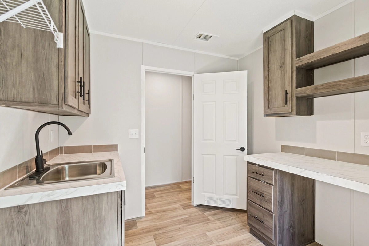 A small, modern utility room with wood cabinets, a stainless steel sink, marble countertops, and open shelving, featuring light wood flooring and a white door.