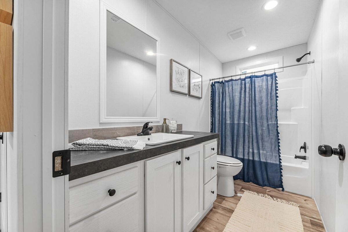 Modern bathroom with white cabinetry, a dark countertop, rectangular mirror, wall art, toilet, and a shower with a blue curtain on a wooden floor.