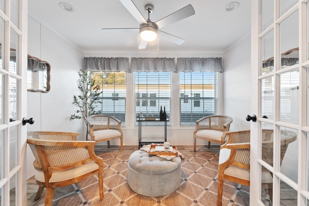 A bright sunroom with four wicker chairs, a round ottoman, a tray with cups, patterned rug, ceiling fan, and three large windows with blinds and gray valances.