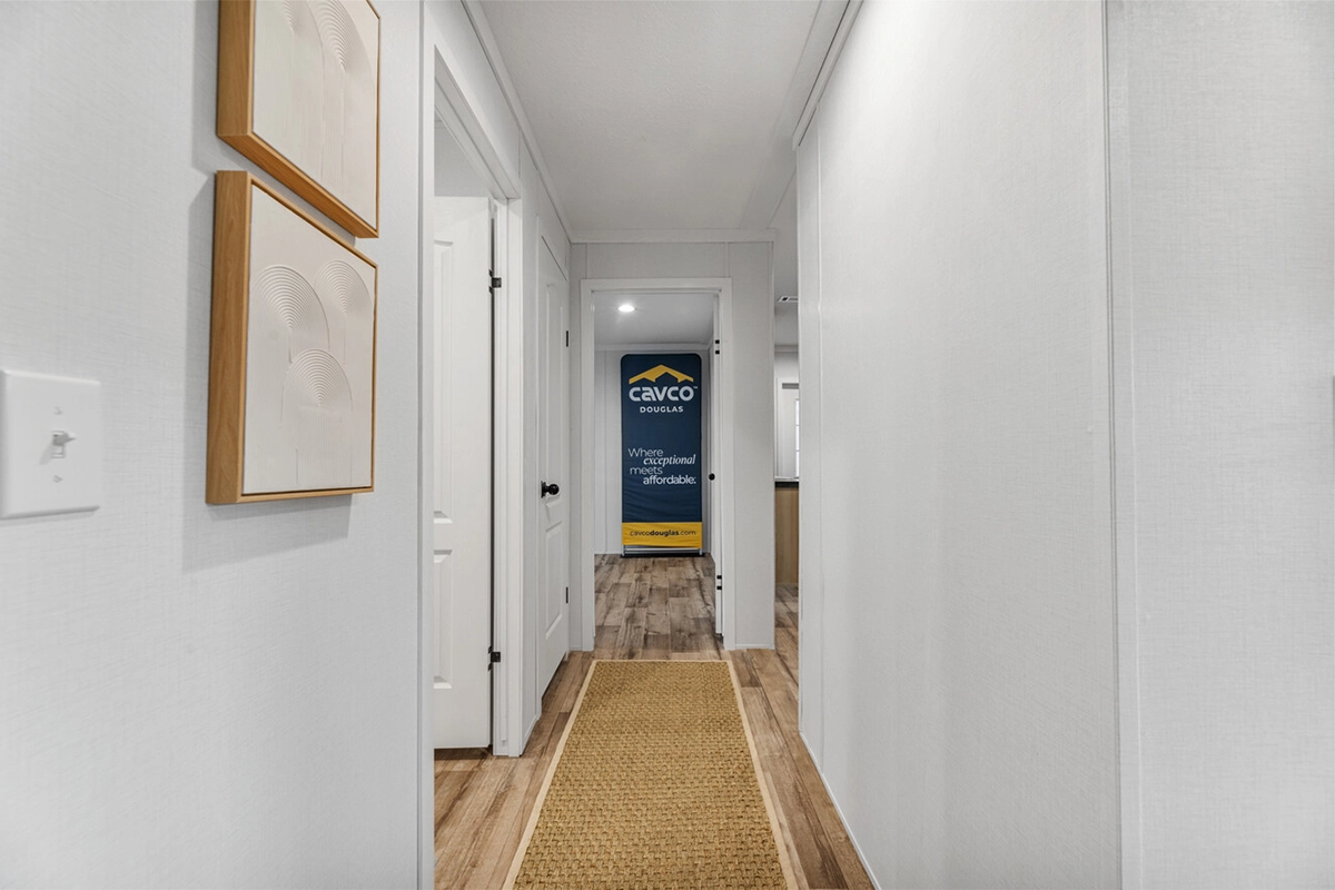 A hallway with white walls, artwork, and a woven rug leads to a room with a blue and yellow Cavco sign visible at the end.