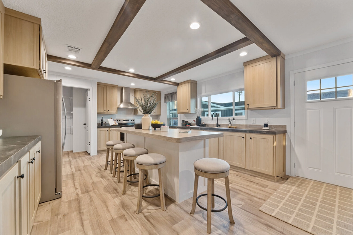 Modern kitchen with light wood cabinets, a large island with four upholstered stools, stainless steel appliances, and a patterned rug by the door.