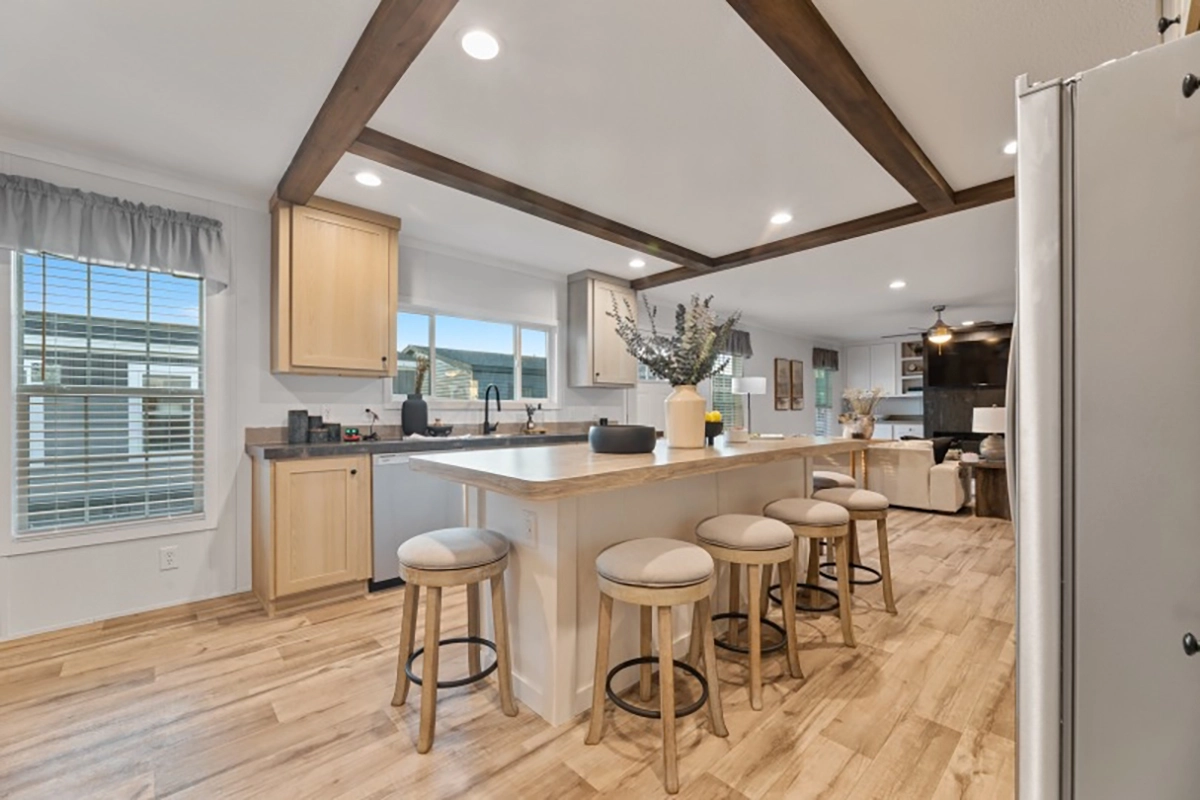 Modern kitchen with light wood flooring, an island with four stools, exposed ceiling beams, and adjacent living area visible in the background.