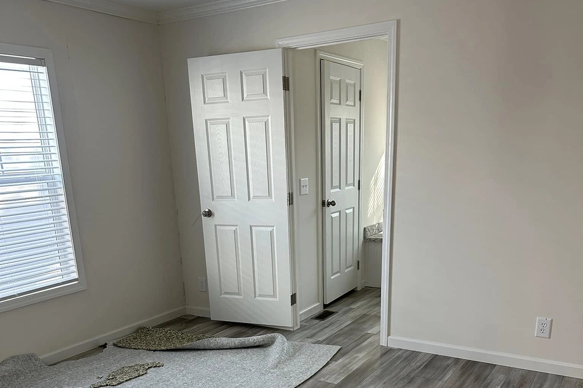 A room with light-colored walls, a window with blinds, two white doors—one open to a small bathroom—and partially removed carpet exposing wood flooring.