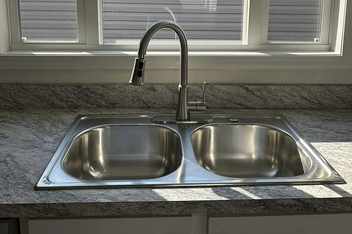 A double-basin stainless steel kitchen sink with a modern faucet, set in a marble countertop beneath a window with blinds.