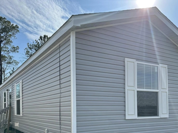 A light gray manufactured home with white trim and shutters, under a sunny sky with trees in the background.