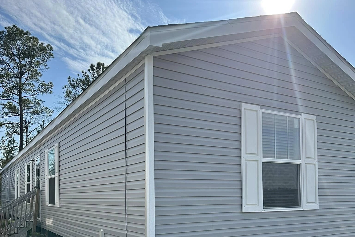 A light gray manufactured home with white trim and shutters, under a sunny sky with trees in the background.