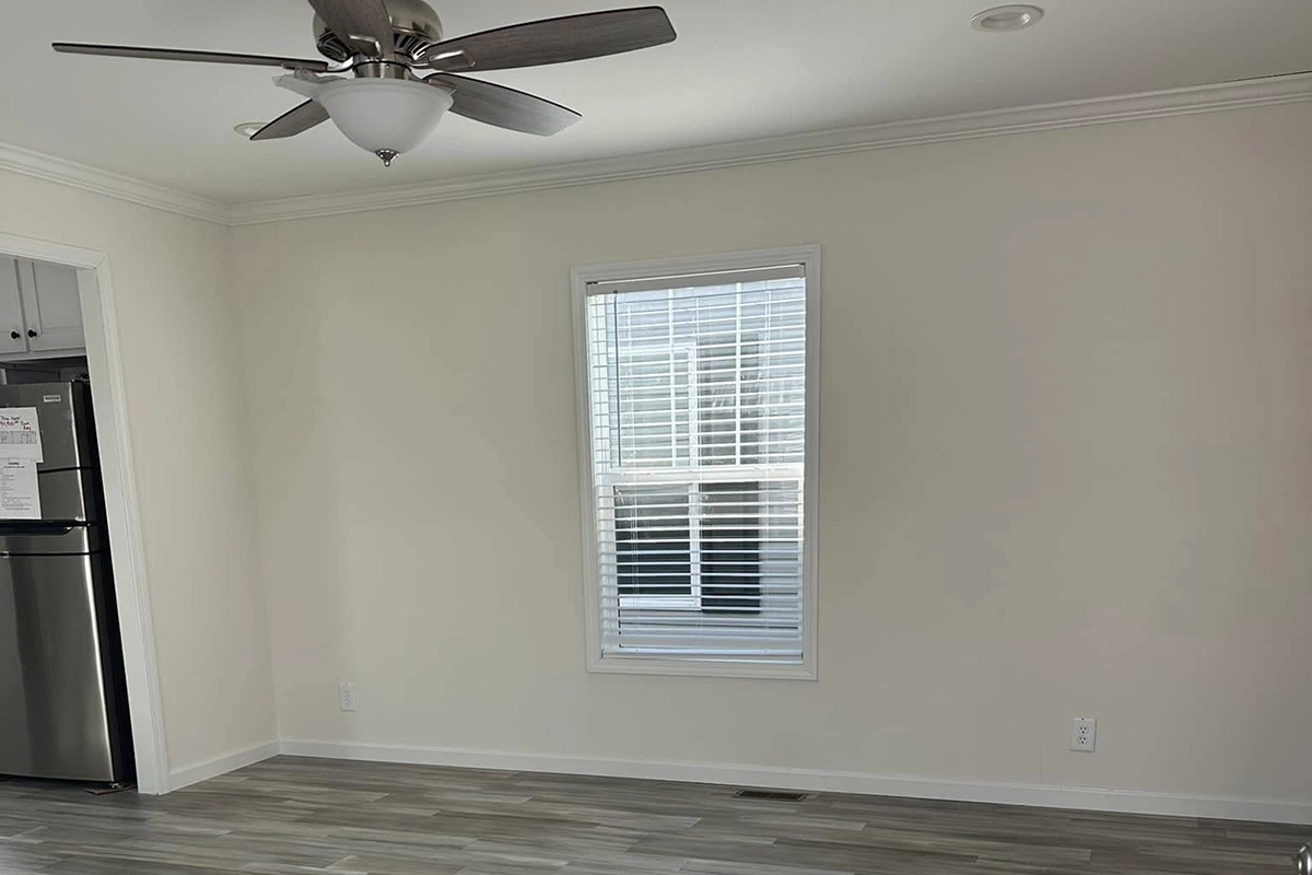 A room with light-colored walls and floor, a ceiling fan with light, a window with blinds, and part of a kitchen with a refrigerator visible on the left.
