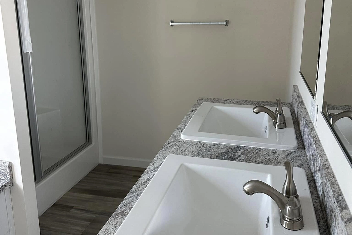 A bathroom with two sinks set in a granite countertop, silver faucets, a large mirror, a towel bar on the wall, and a glass shower enclosure.