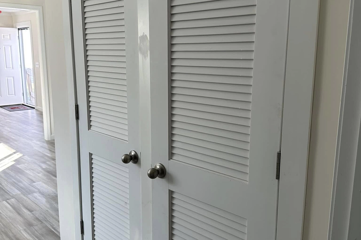 White louvered double doors with round metal knobs in a hallway, with light gray wood flooring and a view of an entry door in the background.
