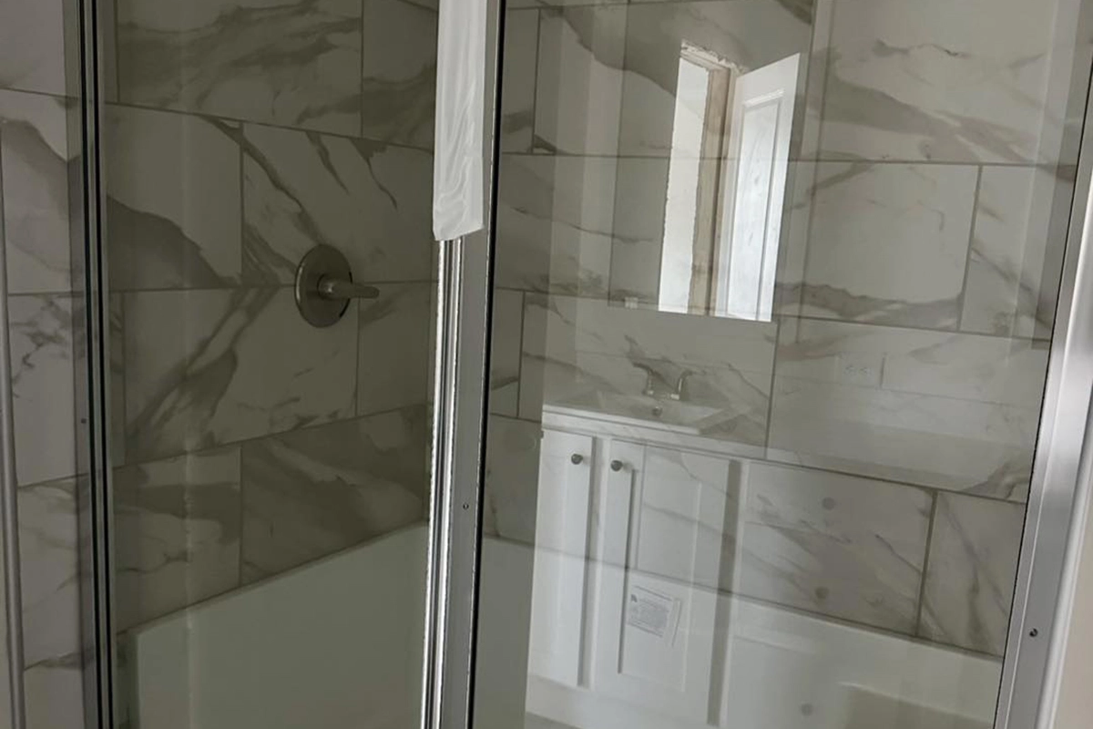 Shower with glass doors and marble-style tile walls, reflecting a white bathroom vanity and sink in the mirror.