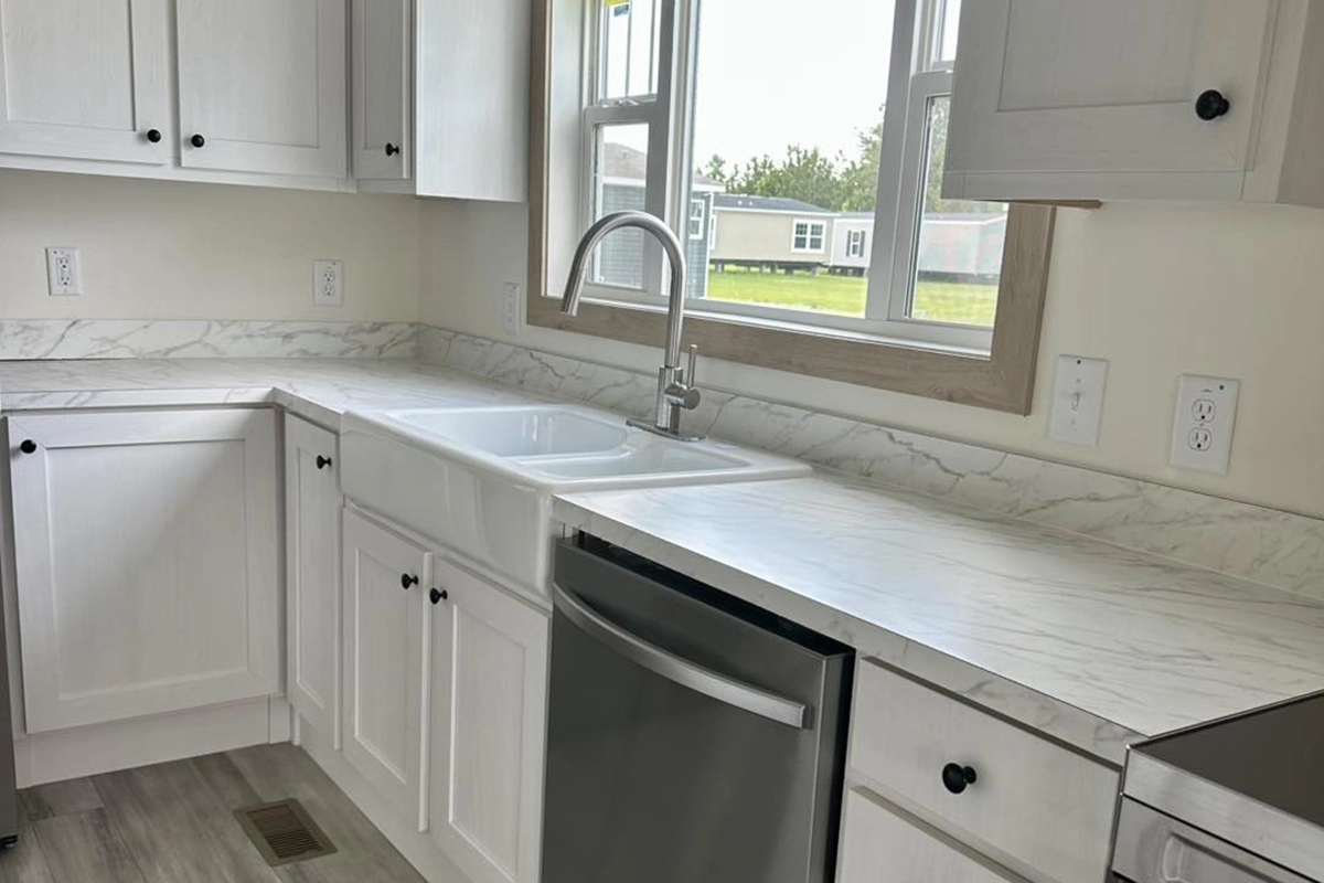 A modern kitchen with white cabinets, marble-patterned countertops, a stainless steel dishwasher, and a sink under a window.