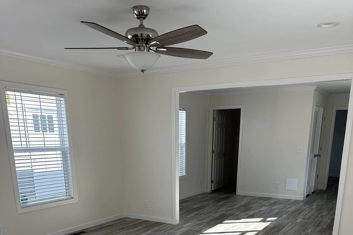 A bright, empty room with light-colored walls, two windows with blinds, a ceiling fan with lights, and wood-patterned flooring. An open doorway leads to another room.