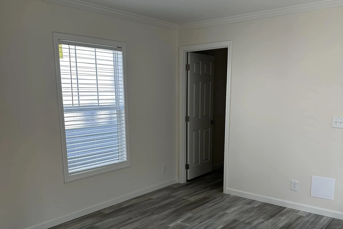 Empty room with light-colored walls, a window with white blinds, wood-look flooring, and an open door leading to another area.