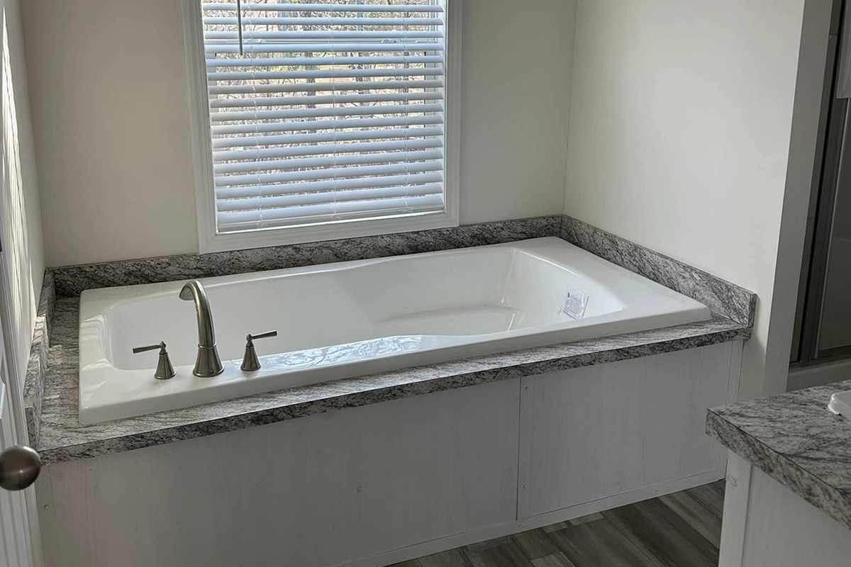 A rectangular bathtub with gray marbled countertop, chrome faucet, and a window with white blinds above it. The walls and bathtub panel are white, and the floor has wood-like tiles.