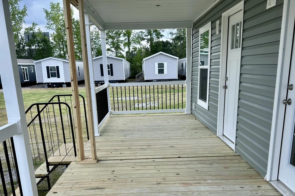 A wooden porch attached to a grey house with white trim, black railing, and a view of several mobile homes in the background.