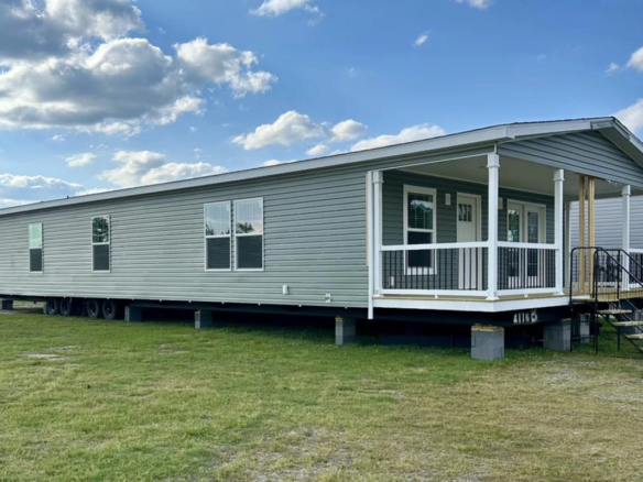 A large, light gray manufactured home with a covered front porch sits on a grassy lot under a partly cloudy sky.