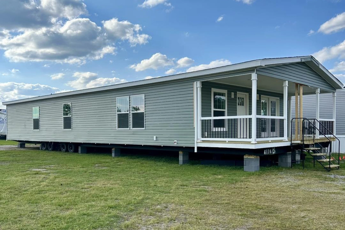 A large, light gray manufactured home with a covered front porch sits on a grassy lot under a partly cloudy sky.