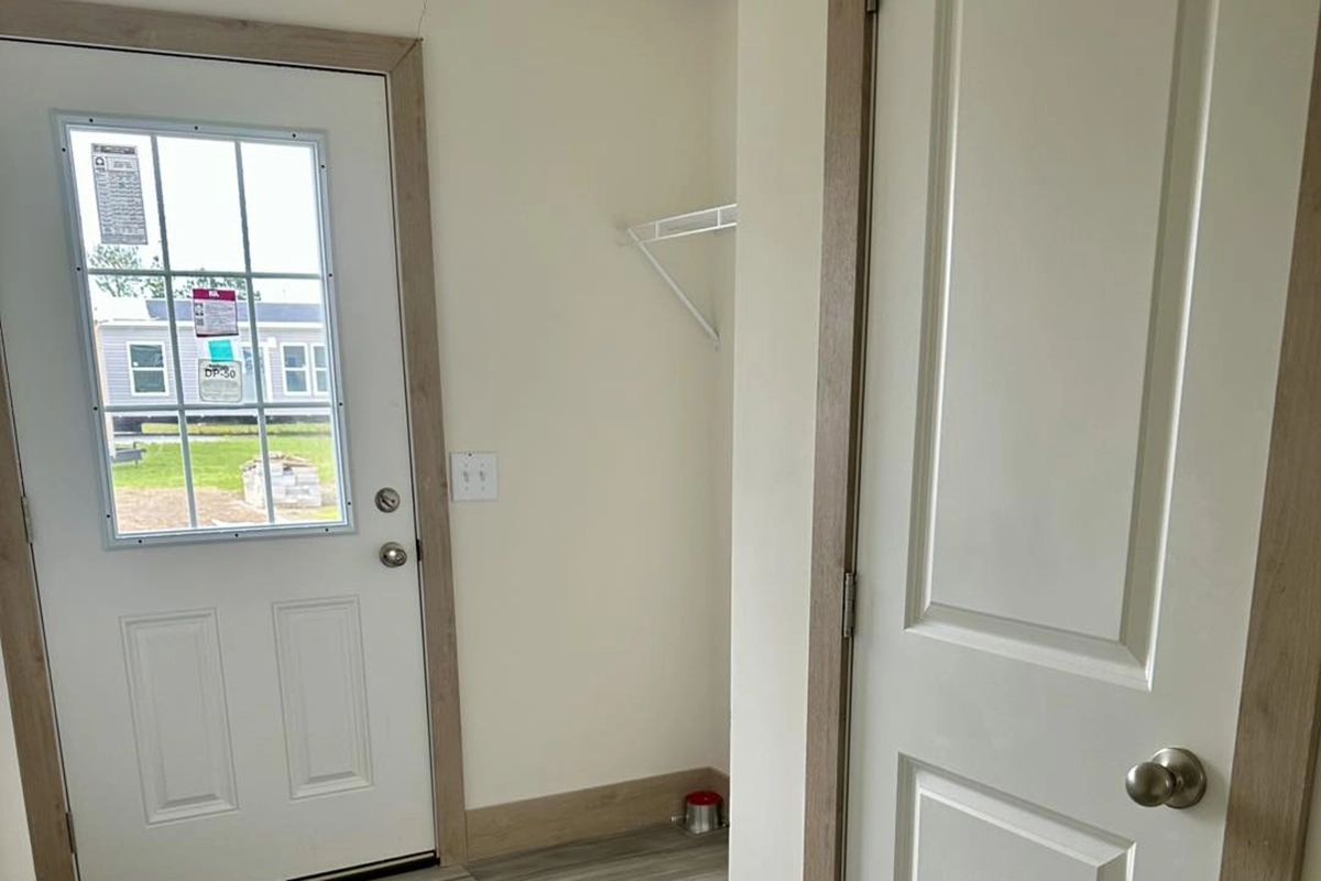 A small room with a white door leading outside, a closet door, a light switch, a wall shelf, and a pet food bowl in the corner.