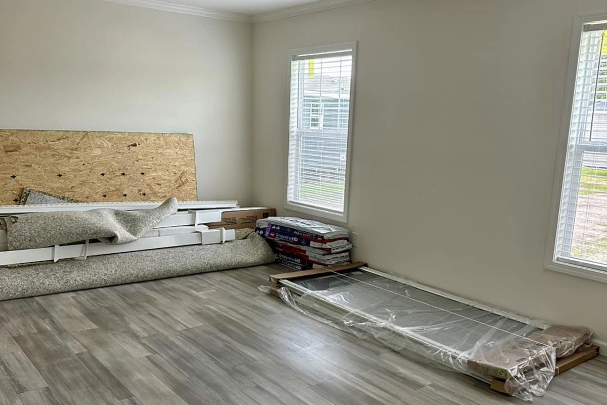 A mostly empty room with light-colored walls and floor, with packaged flooring supplies, doors, and rolled carpet stacked against one wall near two windows.