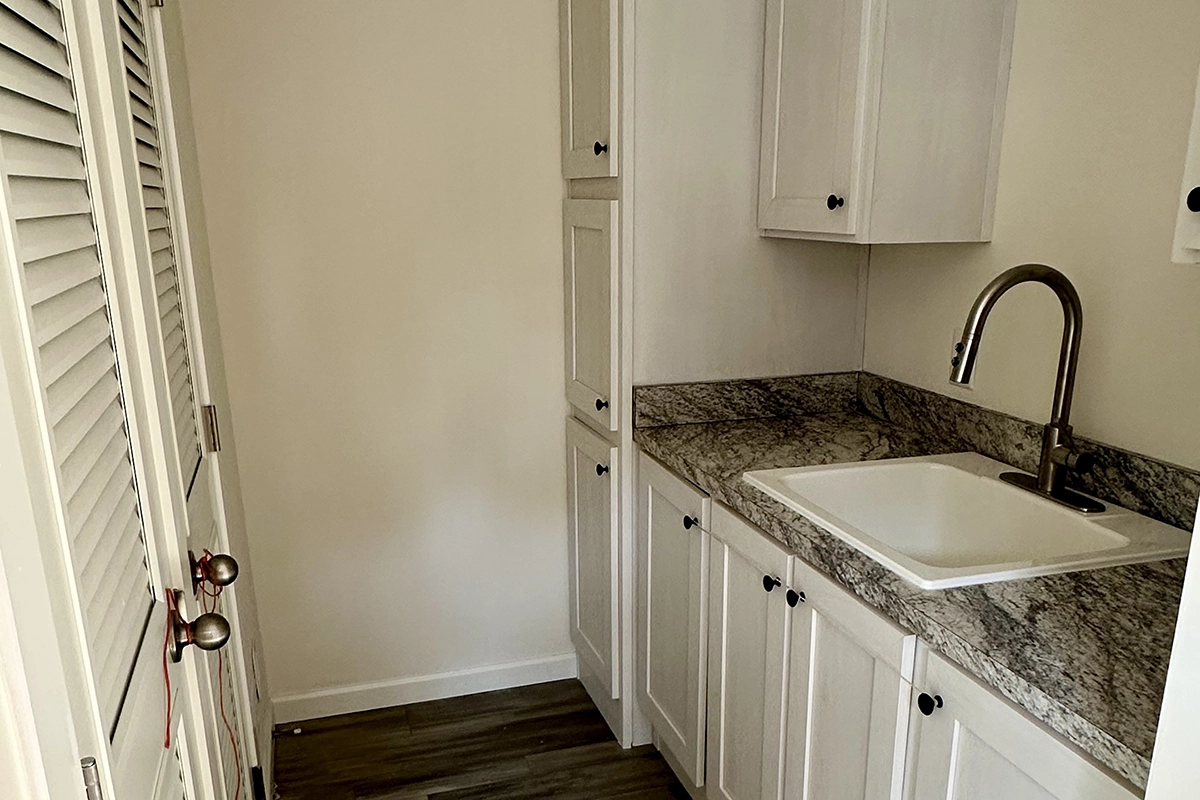 IMG_6384 copy A small laundry room with a marble countertop, white cabinets, a sink with a tall faucet, and louvered closet doors on the left.