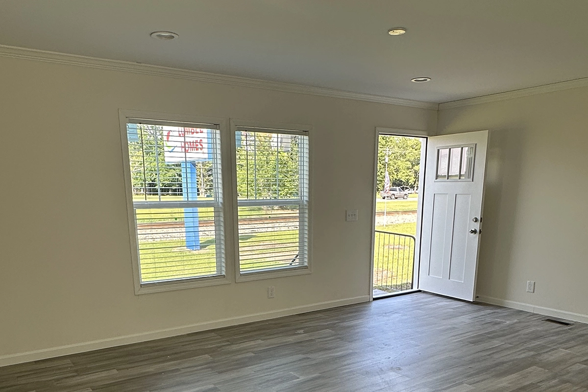 IMG_6387 copy A room with light-colored walls and wood flooring features two windows and an open white door, looking out onto a green lawn and a blue sign visible outside.