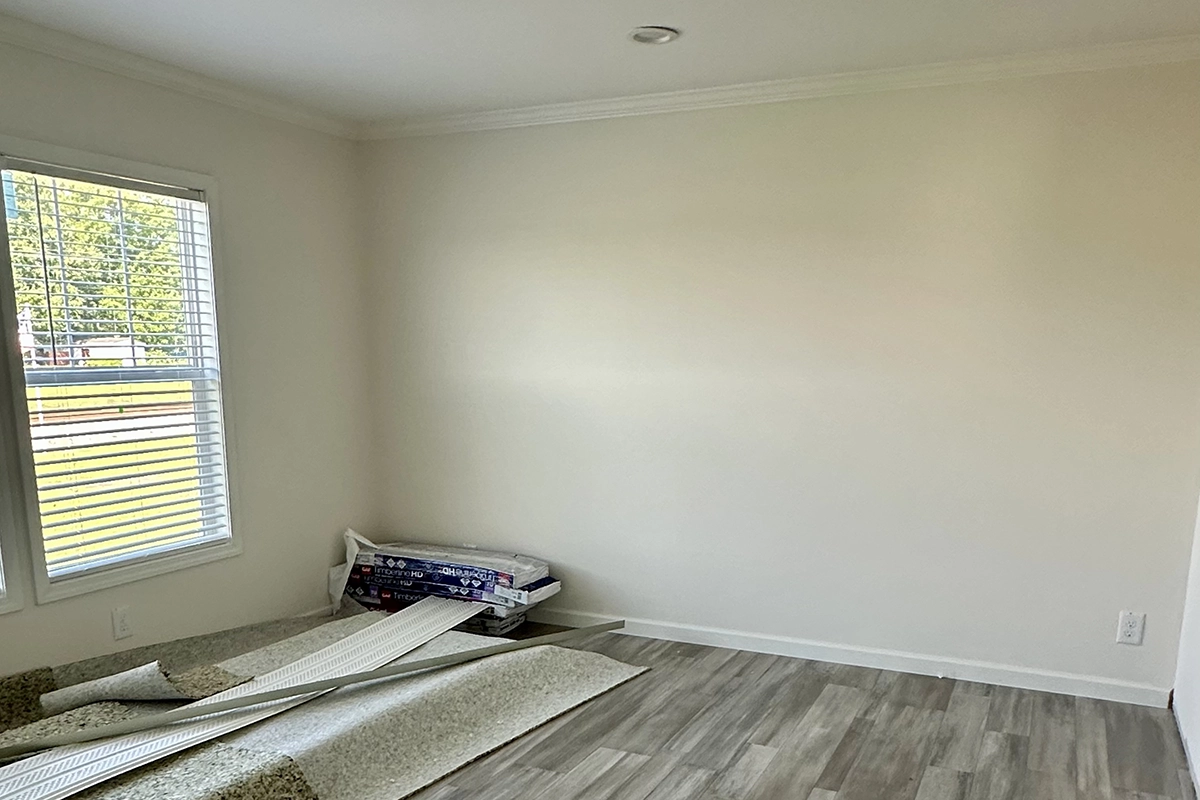 IMG_6389 copy A nearly empty room with light-colored walls, a window with blinds, and flooring being installed; rolls of carpet and floor panels are stacked in the corner.