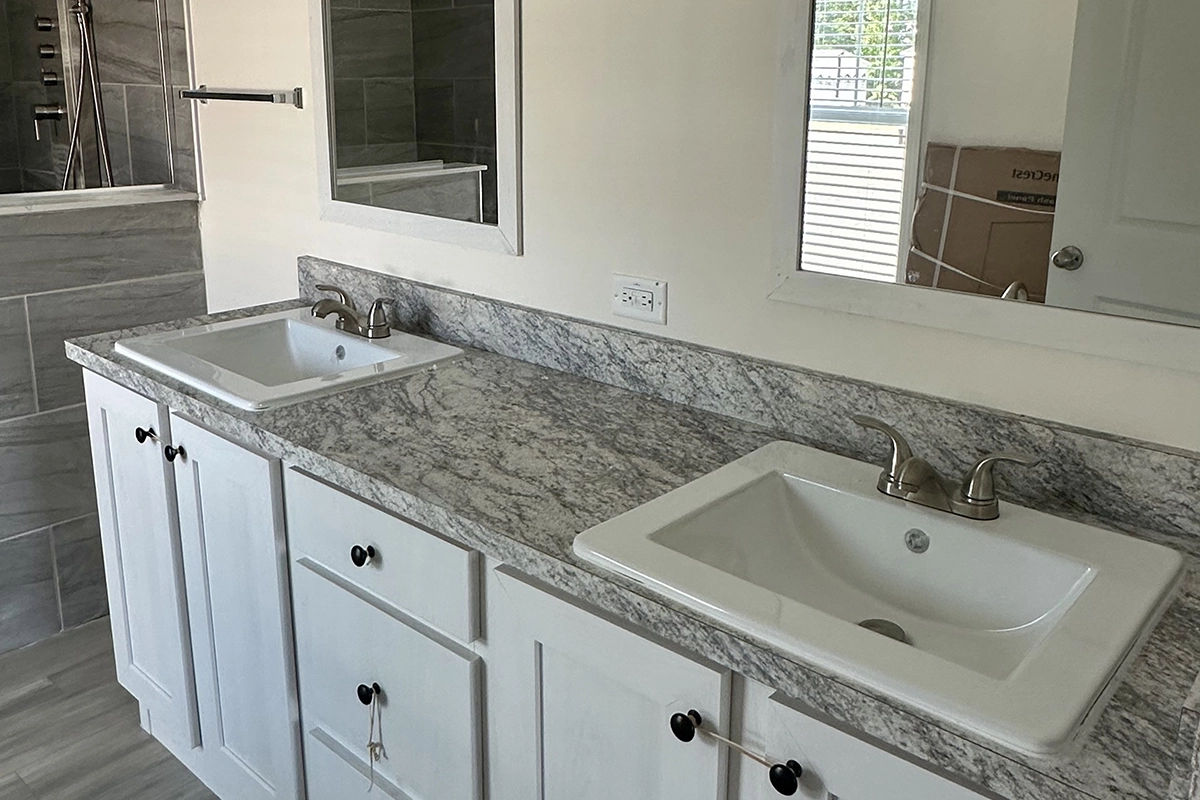 IMG_6390 copy A bathroom with a double sink vanity, marble countertop, white cabinets, wall-mounted mirror, and silver faucets; a shower and window are visible in the background.