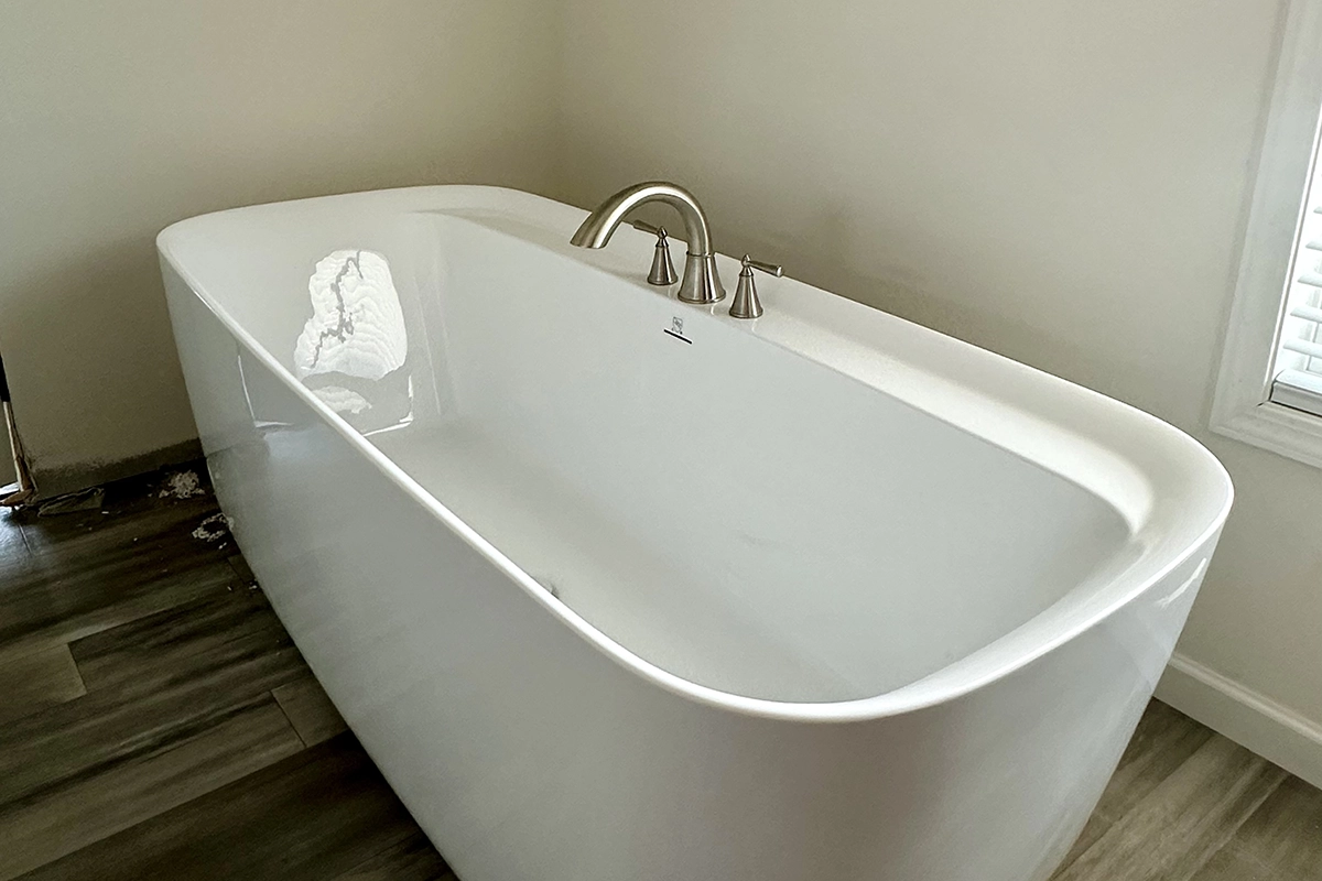 IMG_6391 copy A modern white freestanding bathtub with silver faucet fixtures is positioned next to a window in a bathroom with wood-look tile flooring.