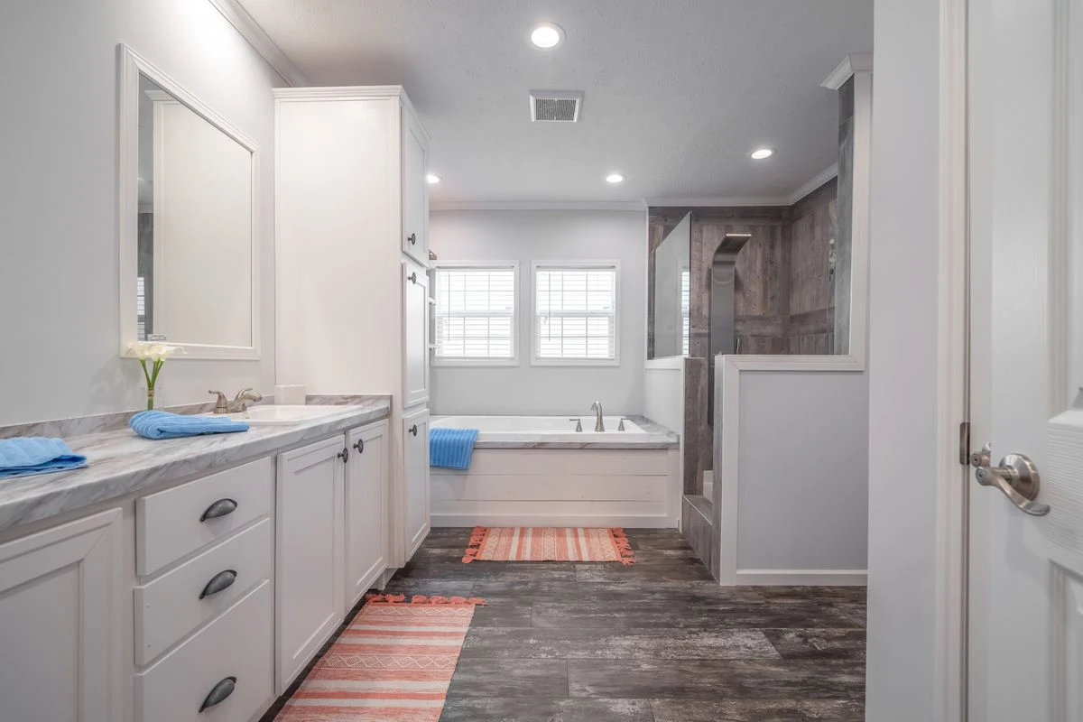 Modern bathroom with white cabinets, double sinks, a built-in bathtub, walk-in shower, two windows, and striped rugs on a dark wood-look floor.