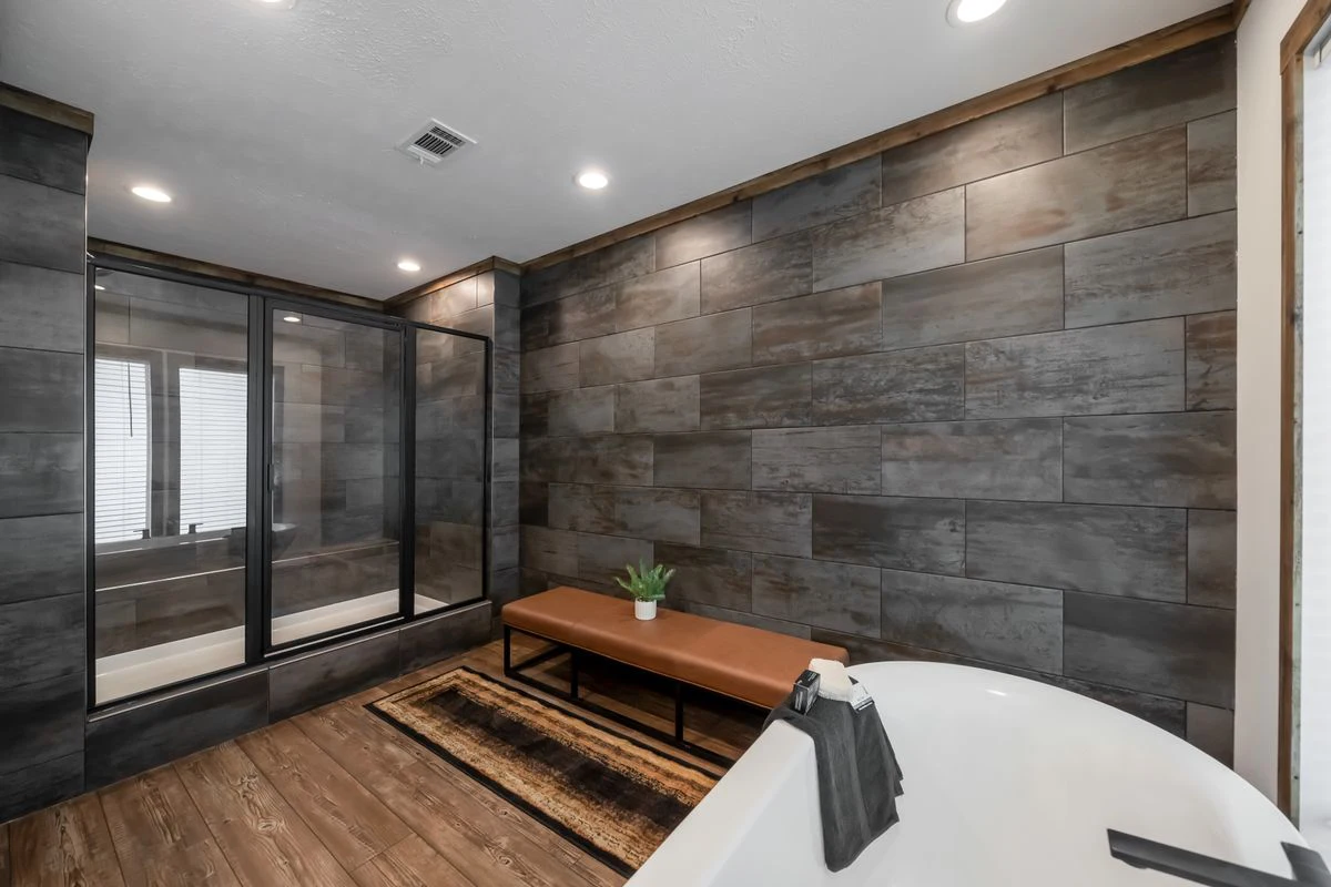 0a4ba240-9016-4b27-b6d0-8f6c3f194d0b Modern bathroom with large dark tiles, a glass-enclosed shower, a brown bench, a rug, wood flooring, and a white bathtub with a towel draped over the side.