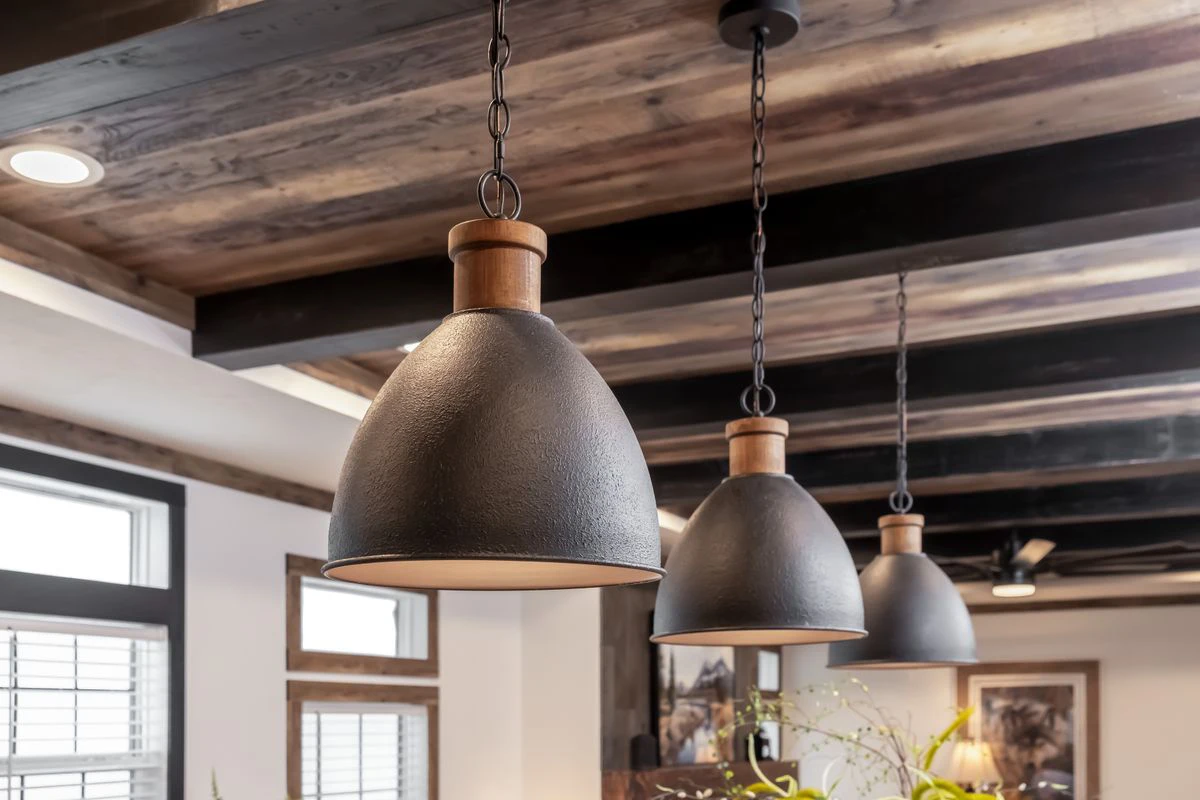 10ae5c2b-a050-482c-b4d7-4f19ce75d321 Three black pendant lights with wooden accents hang from the ceiling of a modern room with exposed wooden beams and large windows.
