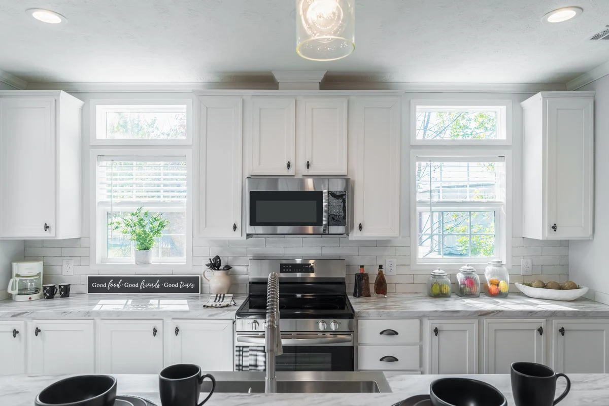 Modern kitchen with white cabinets, stainless steel appliances, marble countertops, a central sink, and jars of fruit on the counter beneath large windows letting in natural light.